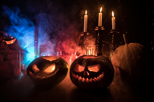 Wizard's Desk. Horror Halloween concept. Magic potions in bottles on wooden table with books and candles. Halloween still-life background with different elements on dark toned foggy background. Selective focus