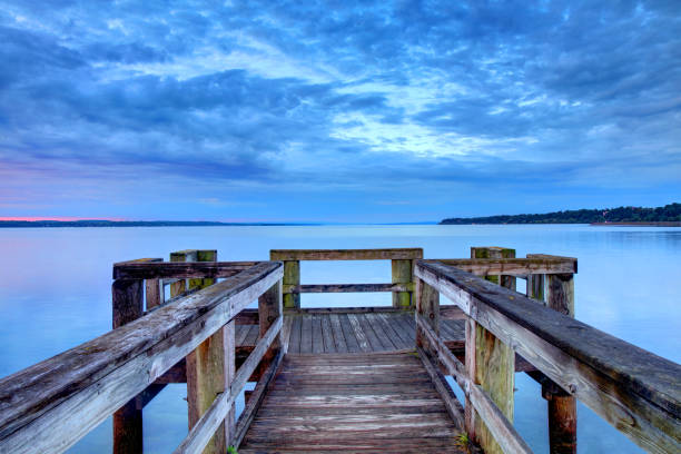 Panoramic view over the water of Penn Cove near Coupeville, WA, USA with a wooden fishing dock and building extended in the water against a blue sky with white feather clouds