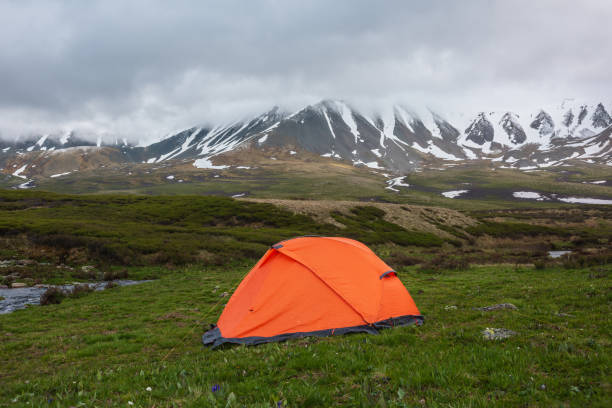istock Orange tent with view to mountain range among low rainy clouds in overcast. Dramatic alpine landscape with snowy mountains in gray low clouds. Bleak atmospheric scenery of tundra under lead gray sky. 1417142129