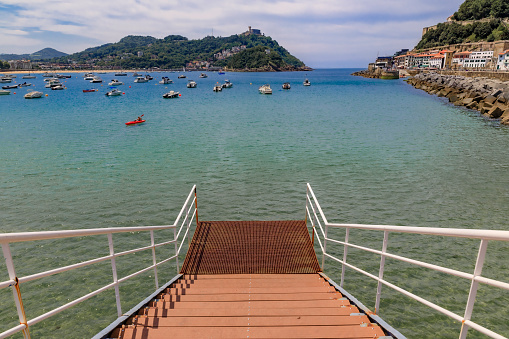 Steps leading into the water in La Concha bay with boats with Santa Clara island in the background, San Sebastian or Donostia in Basque Country, Spain