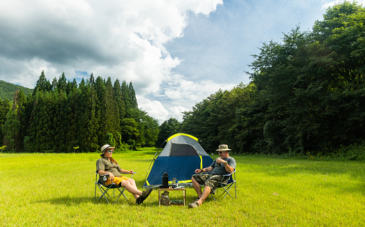 A multi race couple relax in camping chairs by their tent in a wide open lush green grass field surrounded by forest trees on a sunny day.