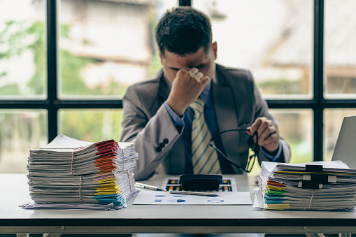 Businessman stressed with work with piles of unresolved documents on desk in office