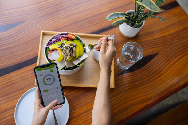 Vegetarian woman eating tofu salad and playing social media. Hipster Asian woman using mobile app checking calories while eating vegan salad cooked by fresh ingredients during morning routine. reduced-calorie diet stock pictures, royalty-free photos & images