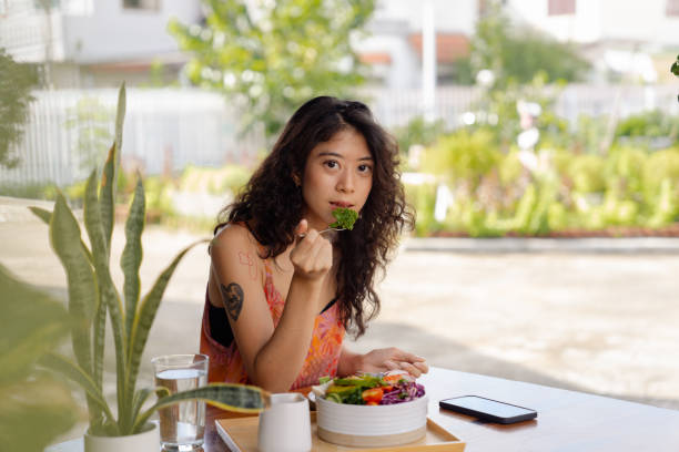 Portrait of woman using smartphone while having plant-based brunch. Smiling Asian woman using smart phone chatting with friends while eating tofu salad a in backyard. image based social media photos stock pictures, royalty-free photos & images