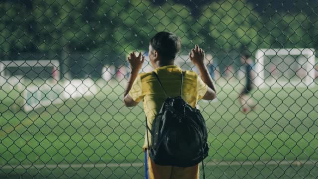 A boy looks into the football field through a chainlink fence