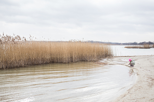 Adorable little girl of 8-9 years old playing by the lake.  
Dressed in a jacket and a pink cap. Winter.  Poland.