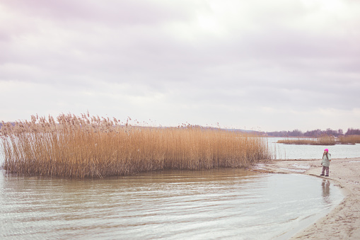 Adorable little girl of 8-9 years old playing by the lake.  
Dressed in a jacket and a pink cap. Winter.  Poland.