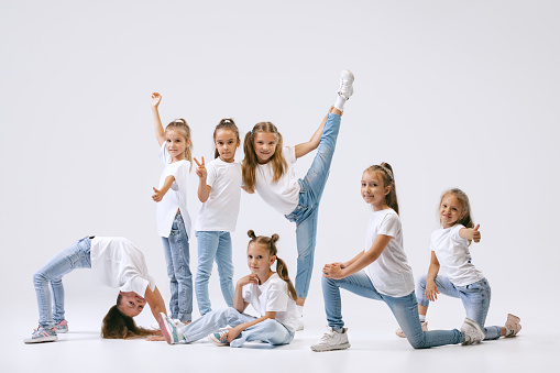 Modern choreography lesson. Dance group of happy, active little girls in jeans and t-shirts dancing isolated on white studio background. Concept of music, fashion, art, childhood, hobby.