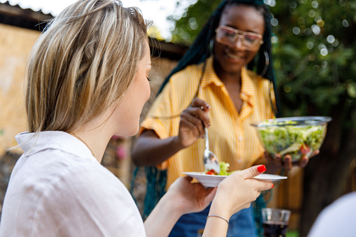 Selective focus shot of smiling young woman sitting at table, holding a plate, while her friend is serving salad for her during a relaxing garden dinner party.