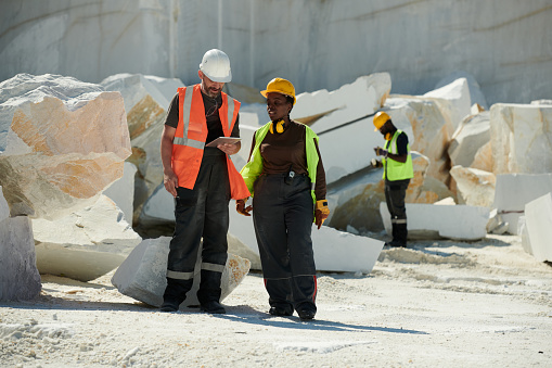 Two young intercultural workers of marble quarry discussing online manual guide while standing against heaps of huge rocks