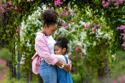 African mother and her daughter hugging each other while happily walking under the blossom trellis arch in the public park during summer for family love and care in mother's day celebration