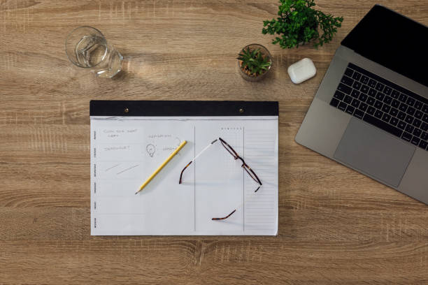A table top view of an array of objects on a desk. There is a laptop, planner pad, eyeglasses and pencil, wireless headphones, glass of water and small plants.