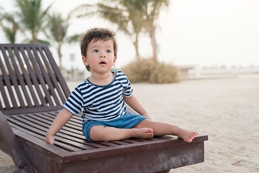Adorable one year old baby boy sitting on the sunbed on the beach enjoying the seaside breeze in an unrecognizable tourist resort