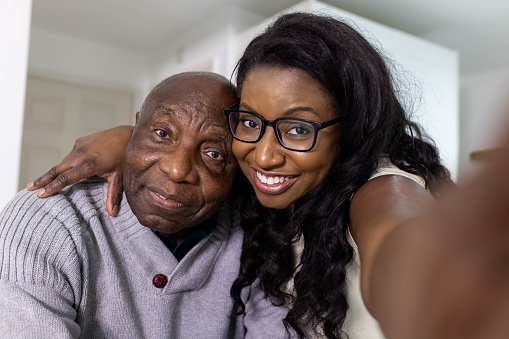A woman takes a selfie with her father who is sitting down. They are both smiling and looking into the camera.