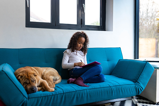 Young girl playing by herself on a digital tablet on a blue sofa. The family pet Shar-Pei dog is asleep next to her.