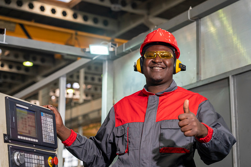 Portrait Of Black Machine Operator In Red Helmet, Safety Goggles, Hearing Protectors And Work Uniform.