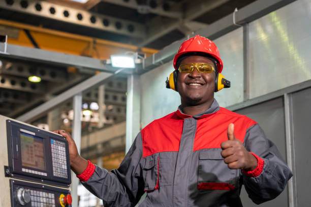 Cheerful African American CNC Machine Operator Standing Next To CNC Controller Console And Giving Thumb Up Portrait Of Black Machine Operator In Red Helmet, Safety Goggles, Hearing Protectors And Work Uniform. metal worker stock pictures, royalty-free photos & images