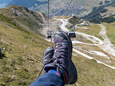 Personal perspective of hiking boots, while sitting on a descending mountain chair lift in the Swiss Alps, with the ground and mountain tracks below. Tele-cabins and open chair lifts are hooked on the line ahead and opposite, as people are transported up and down the steep mountain, during the summer season.