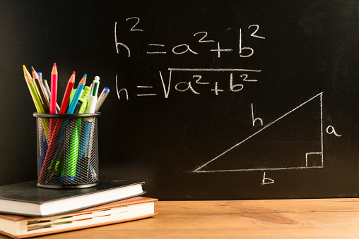 Books, notebooks and a pencil holder with pencils, pens and various school supplies on wooden boards with a blackboard in the background with equations and a triangle written in chalk. Concepts of back to school, education and childhood.