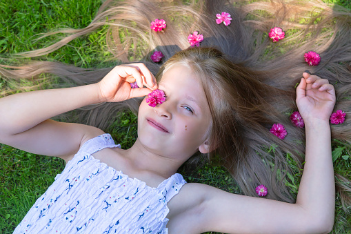 Cute happy caucasian little girl with long loose hair is lying on a grass with purple flowers in her hair in summer