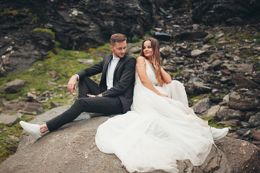 Stylish couple of newlyweds posing sitting on the rocks in the mountains. Attractive couple newlyweds, happy and joyful moment. Wedding travel.
