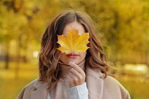 Beautiful smiling woman holding fall yellow maple leaf covering her eyes in autumn park