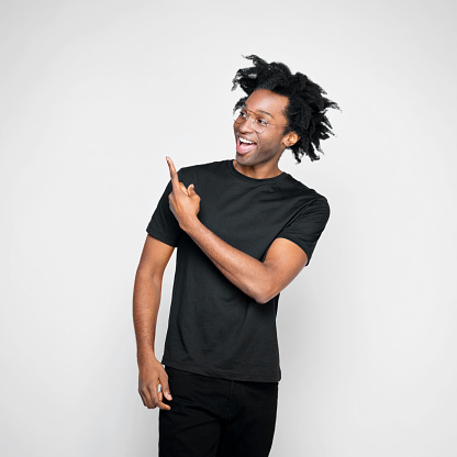 Cheerful afro american young man wearing black clothes, looking away and pointing at copy space. Studio shot on white background.