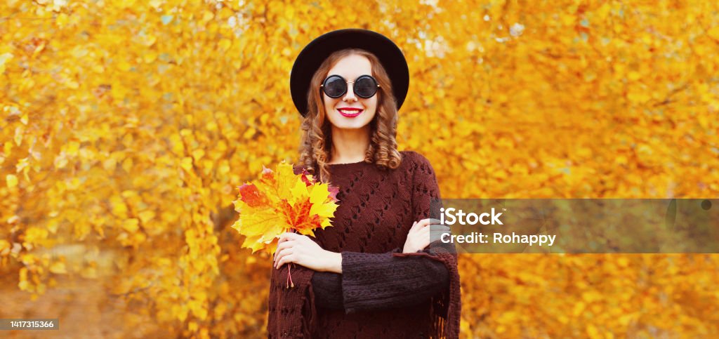 Portrait of stylish young woman with maple leaves wearing black round hat, knitted brown poncho in the park on yellow leaves background 20-29 Years Stock Photo