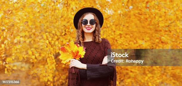 Portrait Of Stylish Young Woman With Maple Leaves Wearing Black Round Hat Knitted Brown Poncho In The Park On Yellow Leaves Background Stock Photo - Download Image Now