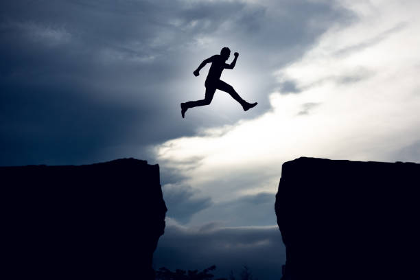 Silhouette young businessman jumping over precipice between two rocky mountains at sunset.