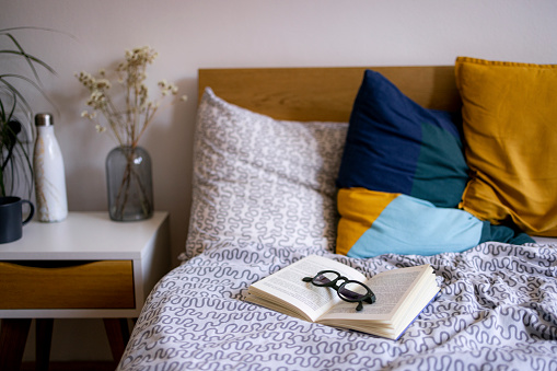 An open book placed on a bed in a bedroom with reading glasses on it, no people