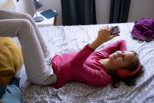 High angle view of a young teenage girl lying on a bed at home in her bedroom and using headphones to listen to the music on her smart phone she is holding in her hands