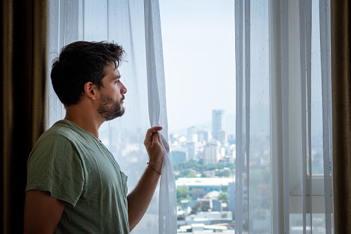 Young man looking out the window of a tall building. Young man looking wistfully outside. Person looking out the window through the curtain