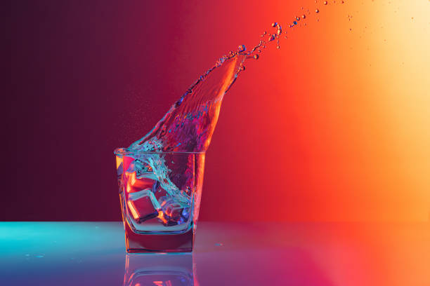 Ice cube falls into a transparent glass of water standing on mirror surface over gradient red orange background in neon light. Art, beauty, drinks Fluid splash. Ice cube falls into a transparent glass of water standing on mirror surface over gradient red orange background in neon light. Concept of art, beauty, drinks. Copy space for ad multi coloured glasses stock pictures, royalty-free photos & images