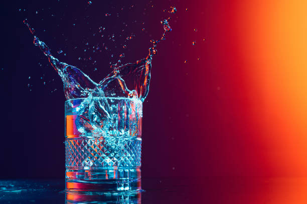 whiskey glass with ice cubes and water splashes standing on mirror surface over gradient black orange background in neon light. art, beauty, drinks - action alcohol alcoholism bar imagens e fotografias de stock