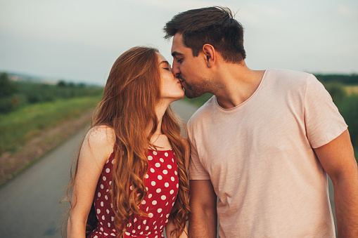 Young caucasian couple in love kissing in the middle of the road in summer evening. Healthy lifestyle. Romantic couple dating. Happy family. Summer road trip. Honeymoon trip. Love, dating.