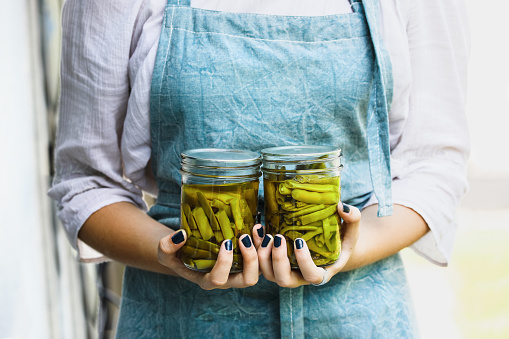 Young woman in a linen apron holding two mason jars filled with fresh home grown green beans. Selective focus blurred background.