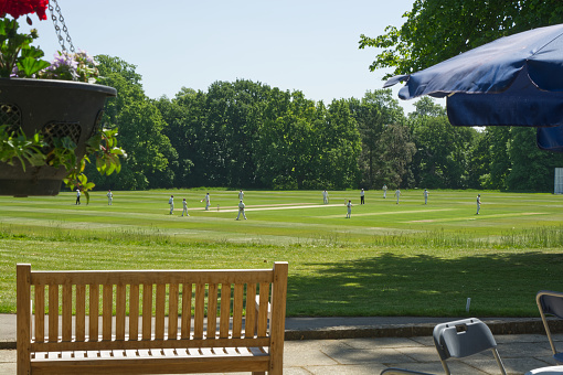 Players on the field during cricket match at Arundel castle ground in West Sussex, England. With patio area and bench in foreground.