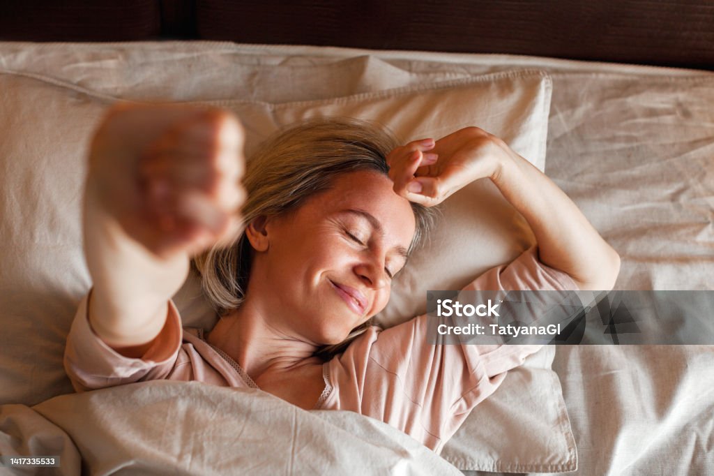 Happy middle aged woman sits on bed and stretching arms Good morning, new day, weekend, holiday. Happy middle aged woman lying on bed, lady stretching arms after sleep and enjoying morning in cozy comfort bedroom interior, free space Sleeping Stock Photo