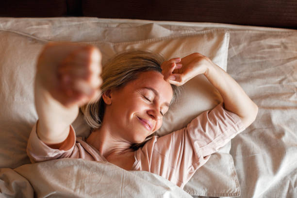 Happy middle aged woman sits on bed and stretching arms Good morning, new day, weekend, holiday. Happy middle aged woman lying on bed, lady stretching arms after sleep and enjoying morning in cozy comfort bedroom interior, free space conscious stock pictures, royalty-free photos & images