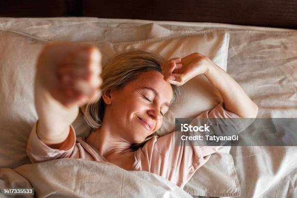 Happy Middle Aged Woman Sits On Bed And Stretching Arms Stock Photo - Download Image Now
