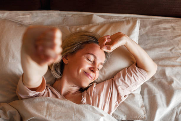 happy middle aged woman sits on bed and stretching arms - woman sleeping imagens e fotografias de stock