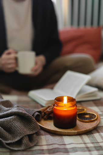 Burning candle on background of young woman holding mug coffee or tea, reading book while sitting in lotus pose on bed in cozy bedroom. Cozy lifestyle, hygge concept