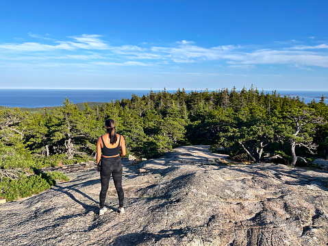 Asian young woman looks over at the forest and seascape below while on top of Cadillac Mountain, the highest point on the eastern seaboard of the USA.