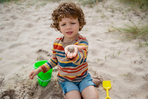 A shot of a young boy sitting down on the sand at a beach in Seahouses, Northumberland. He has been digging in the sand and building sand castles with a beach bucket, he is holding a block of sand in his hand.