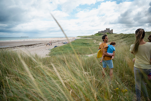 A wide-view shot of a mid adult mother and father walking to the beach with their young children in Seahouses, Northumberland. The father is carrying a bodyboard.