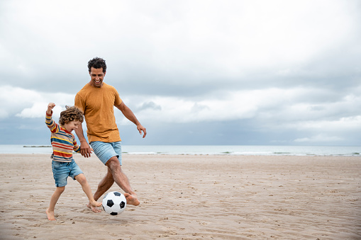 A shot of a mid adult father and his young boy playing football on the sand at a beach in Seahouses, Northumberland. They are having a fun time together.