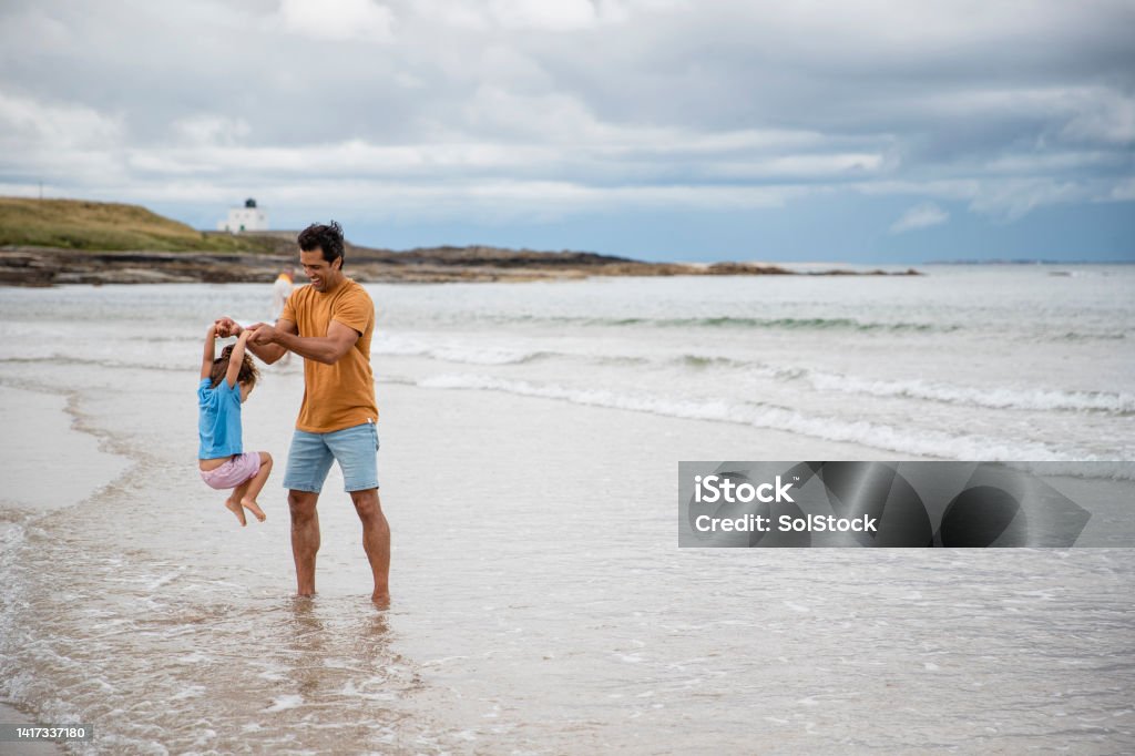 Don't Drop Me! A shot of a mid adult father carrying his young daughter while standing on the sand at a beach in Seahouses, Northumberland. He is holding her above the water from the tide and she doesn't want to get her feet wet. 2-3 Years Stock Photo