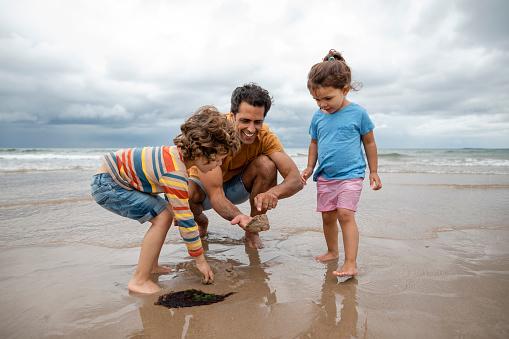 A shot of a Hispanic family standing on the sand at a beach in Seahouses, Northumberland. They are exploring the beach, the father is crouching and holding a handful of sand with an insect in his hand.
