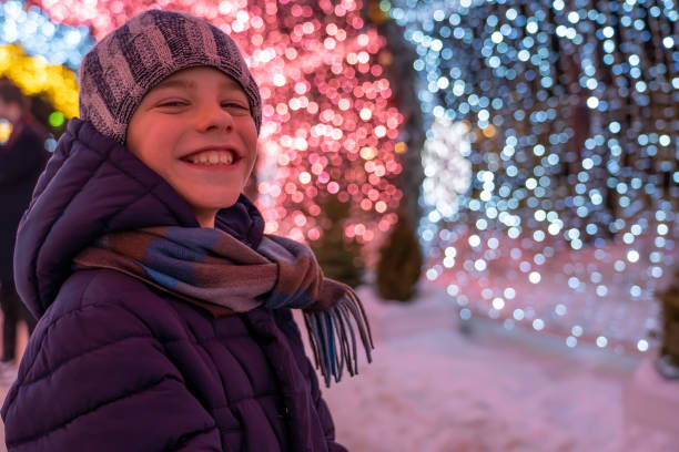Teen boy smile in warm clothes against background of glowing Christmas decorations at winter. Festive garland lights. stock photo